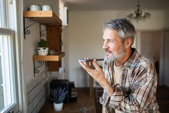 Middle-aged man using voice assistant on smartphone in home kitchen