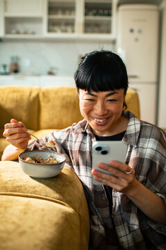 Smiling woman eating breakfast while using smartphone at home