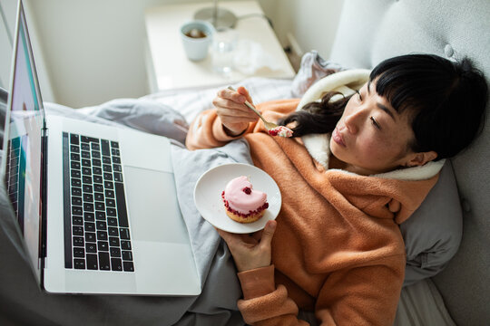 Woman relaxing in bed with laptop and cupcake at home