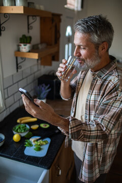 Mature man drinking water and checking smartphone in home kitchen