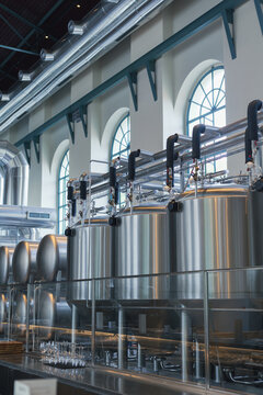 Modern brewery interior with stainless steel fermentation tanks and industrial brewing equipment inside a historic building with large arched windows. Professional beer production facility combining