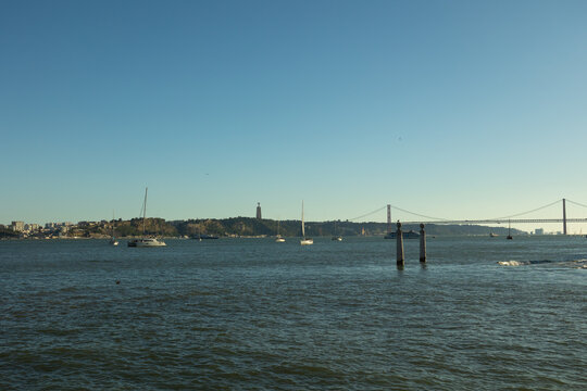 Panoramic view of the Tagus River with the 25 de Abril Bridge and the Cristo Rei monument in Lisbon, Portugal. Sailboats resting on calm waters under a clear blue sky create a peaceful waterfront