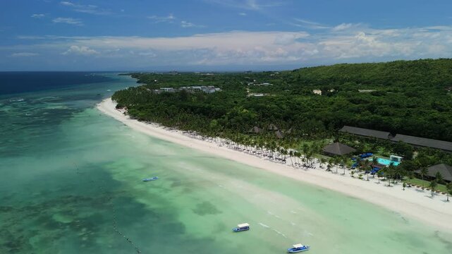 Aerial ascends and pulls back from Dumaluan Beach, revealing white sand, turquoise shallows, boats, palm-lined resorts, and the coastline of Panglao, Bohol