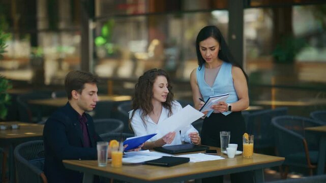 Business professionals engage in a collaborative outdoor meeting at a cafe. They are discussing documents and taking notes in a professional setting.
