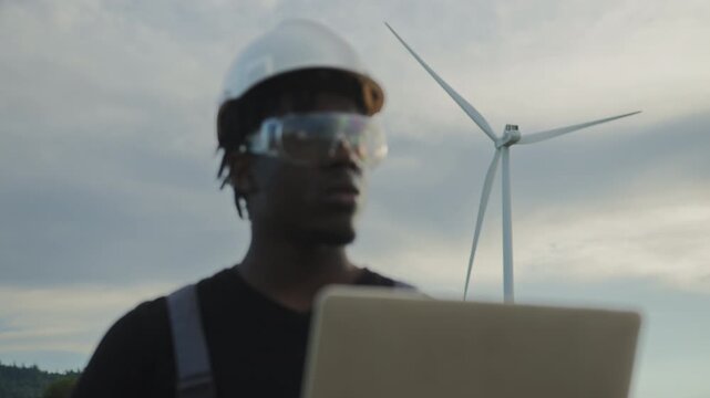 Man in hard hat and safety glasses uses laptop near wind turbine in field, monitoring renewable energy production. Clean electricity, eco-friendly technology, sustainable industry, future innovation