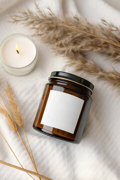 Amber glass jar mockup with blank white square label on textile background with pampas grass and burning candle.