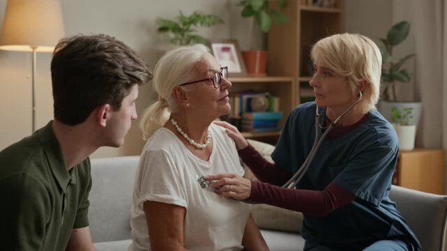 Middle aged Caucasian female nurse using stethoscope to examine senior Caucasian woman sitting next to youngadult son. Video is perfect for promoting home healthcare services