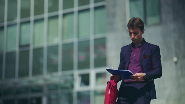 A young professional man reviews documents outdoors in front of a modern office building. He appears focused and diligent, preparing for or reflecting on an important business task.