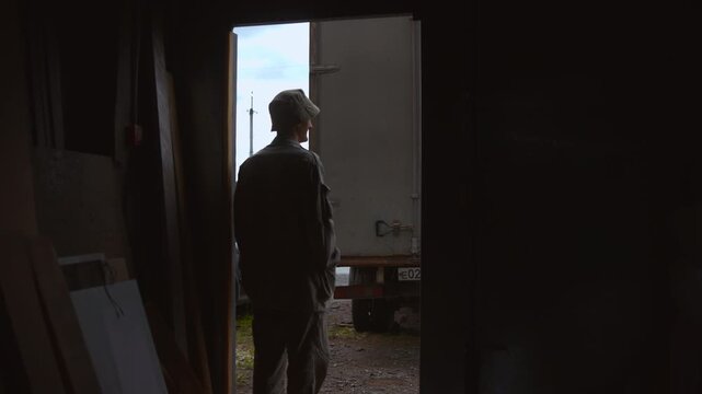 Worker stands in doorway watching parked box truck from dark workshop interior, backlit silhouette framed by rough opening with industrial textures and quiet waiting mood