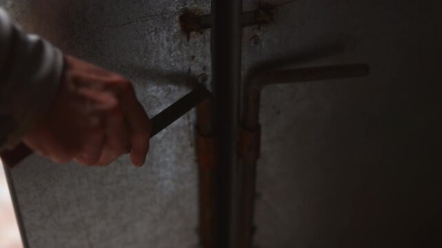 Rusty metal latch pulled outward by hand inside dim industrial enclosure with textured galvanized panel, showing worn hardware, lateral movement, corrosion, grip detail, and utilitarian surface aging