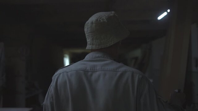 Rear view adult in light hat and loose jacket pausing inside dim workshop with cluttered storage, fluorescent rim light, shadowed face, and industrial textures, with person, adult, hat, jacket detail