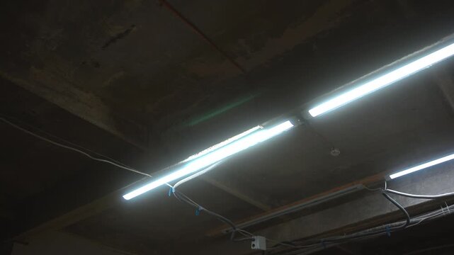 Low angle industrial ceiling with illuminated fluorescent tubes, exposed conduit, junction hardware, and rough unfinished concrete texture, with fluorescent, light, ceiling, concrete detail