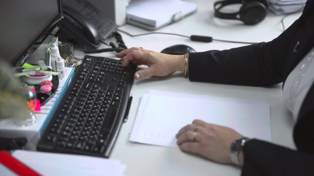 Office worker hands typing on keyboard while steadying stack of documents beside mouse, telephone, headset, and desk accessories in bright workspace close view, with keyboard, office, hand, document