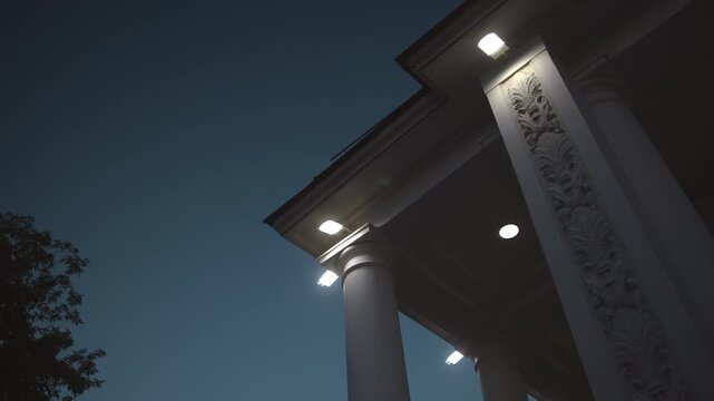 Low-angle evening view of classical portico with illuminated roofline, ornate carved relief, smooth columns, and cool-white fixtures beneath grand entablature, with architecture, portico, column