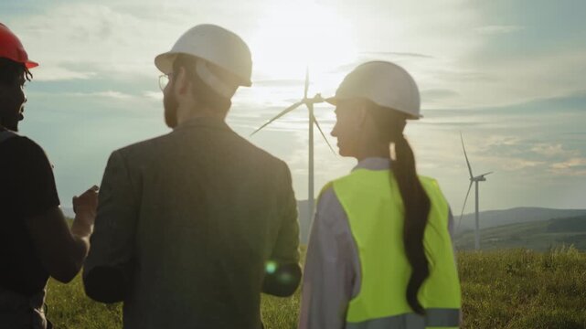 Engineers in hard hats and safety vests converse outdoors amidst rolling hills and wind turbines at sunset. Clean electricity, eco-friendly technology, sustainable industry, teamwork, collaboration