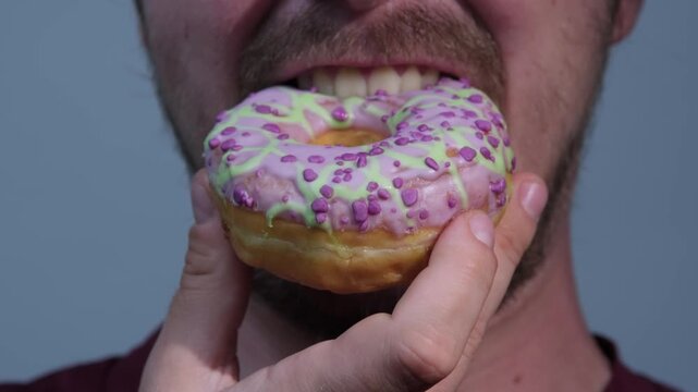 Man holds colorful donut close to mouth in a simple setting and eats it