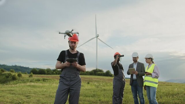 Technician in hard hat and overalls controls drone with remote in grassy field with wind turbines under cloudy sky. Clean electricity, eco-friendly technology, sustainable industry, professional