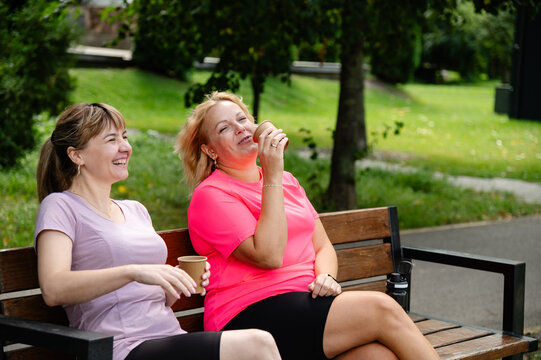 Two women sitting on a bench in a park sharing drinks and laughter on a sunny day