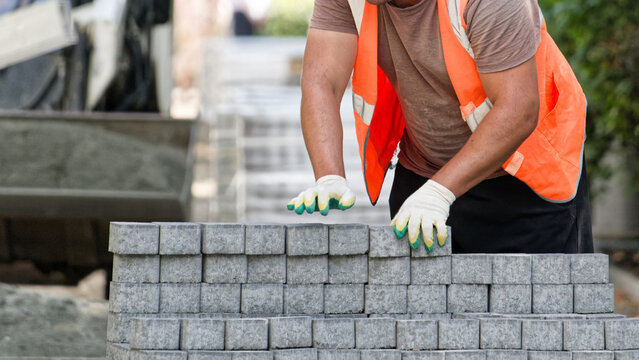 Close-up of a laborer arranging gray paver blocks by hand at a construction site, showing manual work, masonry materials, and paving preparation. Photo