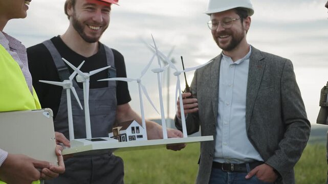 Engineers discuss wind turbine model and tablet in field with windmills. Their teamwork emphasizes renewable energy planning, clean electricity, and sustainable technology for modern industry