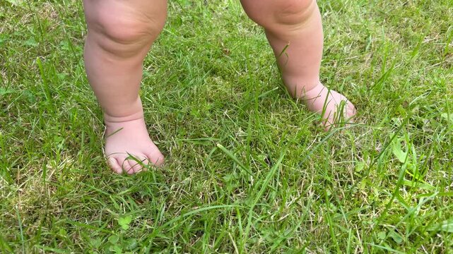 Close-up of a baby's bare feet standing and squatting on green grass in a diaper. Early childhood exploration and play outdoors