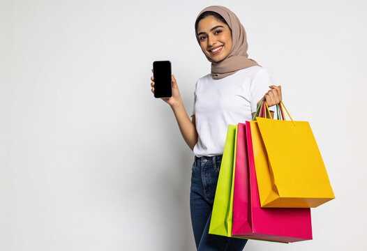 Smiling Muslim woman in hijab holding smartphone with blank screen and colorful shopping bags on white background