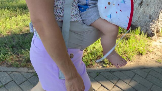 a mother walking in a sunny park while carrying her baby in a front-facing gray mesh baby carrier. Modern parenting and family outdoor activities