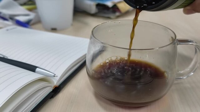 Pouring hot black coffee into clear glass cup on office desk next to open notebook and pen