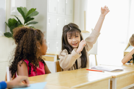 Smiling schoolgirl raising her hand during a diverse classroom lesson, showing active learning, student participation, confidence, friendship, education, teamwork, and inclusive childhood development.
