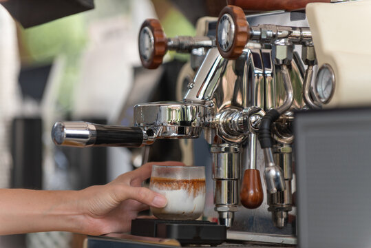 Hand holding glass of milk under coffee maker for espresso extraction, authentic photo, not AI.