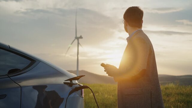 Man in suit jacket connects charging cable to electric car near wind turbines at sunset. The moment highlights renewable energy, clean electricity, and sustainable technology for modern mobility.