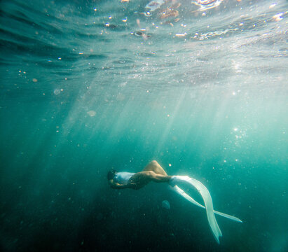 Underwater freediving, woman swimming with long white fins in clear turquoise sea surrounded by sunbeams