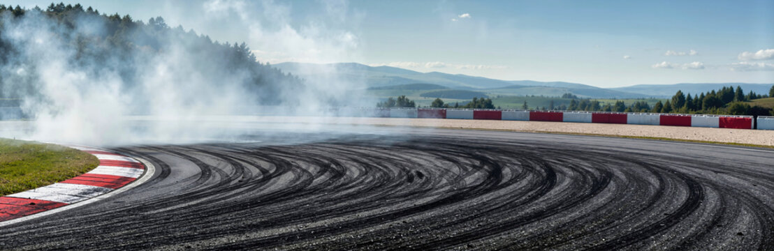 Tire smoke and black skid marks on asphalt race track curve during drift action. Curved speedway road with red and white curb barrier against mountain landscape under daylight, motorsport banner