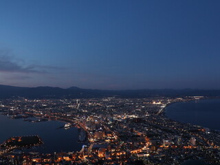 Fototapeta premium Coastal Cityscape at Dusk with Harbor Lights – Calm Sea Reflection and Mountain Silhouette in Evening Glow