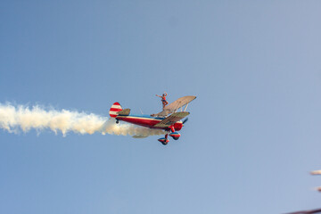 Wing walker standing on top of a red, white, and blue biplane during an airshow with white smoke trail © hcarbuccia
