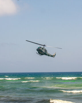 Military utility helicopter flying low over ocean waves near the coastline under a clear sky