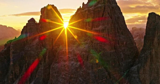 Aerial drone flight toward the glowing sunset between the iconic Tre Cime peaks in the Italian Dolomites. Dramatic alpine landscape with golden sunstar, lens flare and towering mountain cliffs.