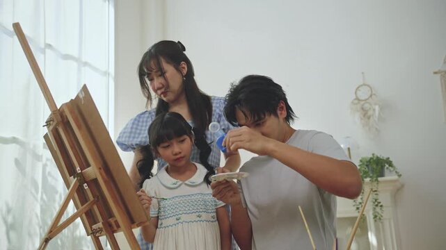 Asian family enjoying painting together at home. father, mother, and young daughter doing creative art activity with paint and canvas in a bright living room, creativity, and learning through play.