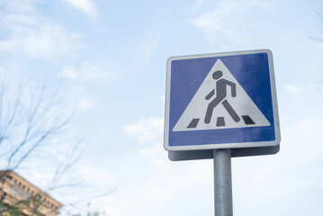 A pedestrian crossing sign stands against a clear blue sky. The sign features a triangular shape with a walking figure, indicating a safe crossing area. © Ksu