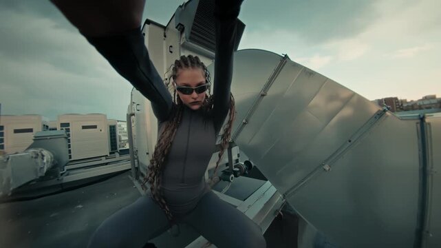 Confident young woman balancing on roof edge with arms spread beside industrial vents and bright sky