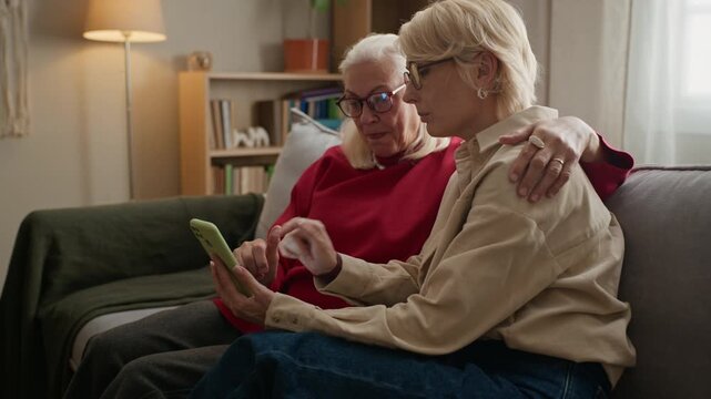 Senior Caucasian woman and middle aged daughter sitting together, using smartphone and smiling. Video represents senior care, family connection, and digital literacy for elderly