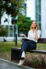 Fototapeta premium Young businesswoman sitting on a park bench talking on a smartphone and holding a tablet