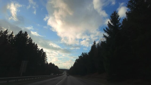 Car journey POV driving on a scenic highway through a dense forest under a dramatic cloudy sky during sunset.