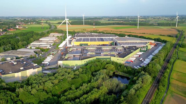 Aerial drone footage of the Colruyt distribution center in Halle, Belgium, showcasing sustainable energy with wind turbines and solar panels at sunset.