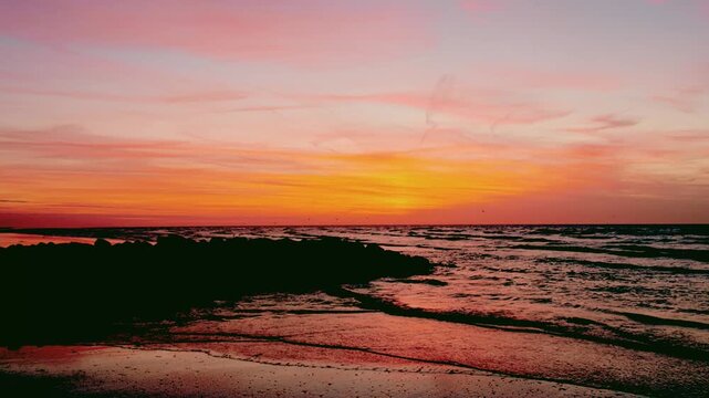 Vibrant Pink and Orange Sunset Over the Ocean with Gentle Waves Lapping on a Rocky Shoreline. Cinematic and Serene Seascape.