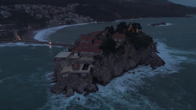 Drone Diagonal Pull-Away from Sveti Stefan Island with Evening Lights and Crashing Sea Waves in Overcast Weather