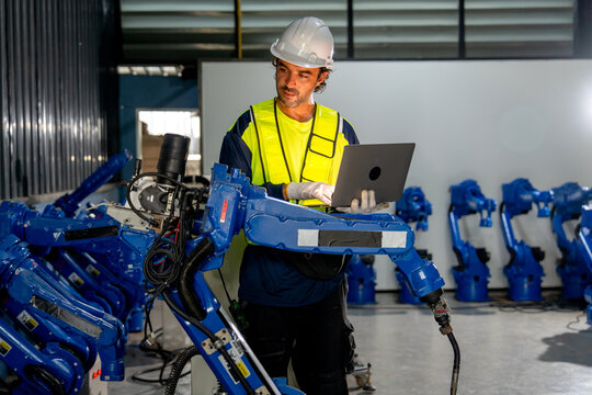 Skilled technician in yellow vest uses laptop to program blue robotic arm in industrial workshop with corrugated metal walls
