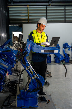 Engineer in safety gear programs blue robotic arm with laptop in industrial workshop with corrugated metal walls