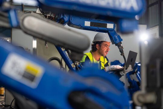 Engineer in high-visibility vest programs blue robotic arm using laptop in industrial workshop with corrugated metal walls