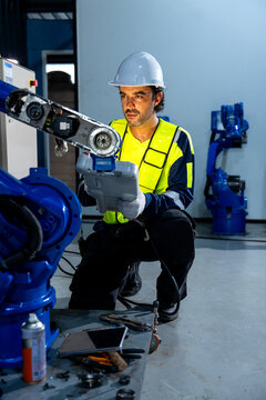 Engineer in yellow vest and white helmet adjusts blue robotic arm with focused intensity in clean industrial workshop